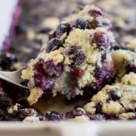 A spoonful of fresh-baked blueberry cobbler being scooped from a white baking dish full of bubbling blueberries.