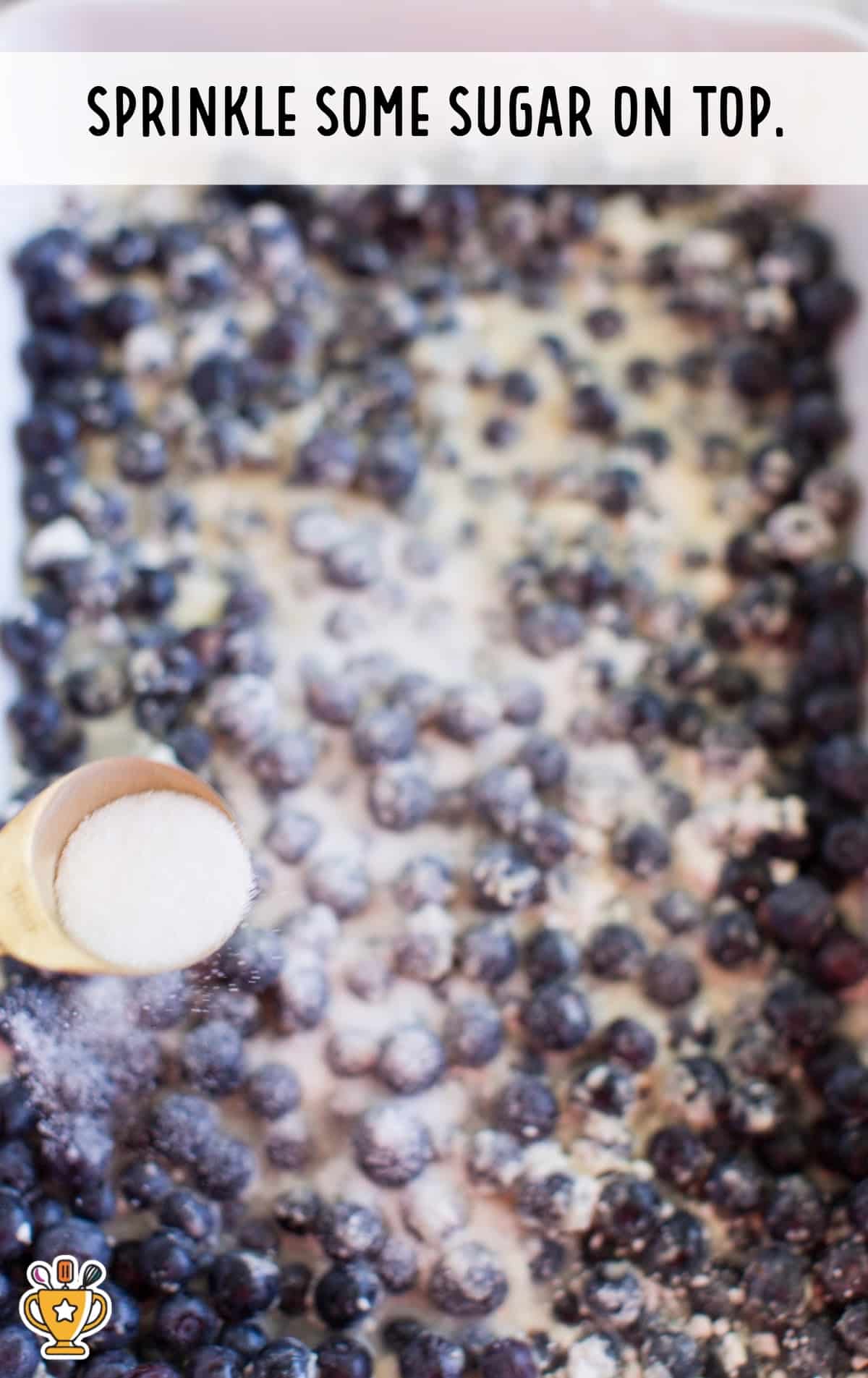 Granulated sugar being sprinkled from a gold measuring spoon over a layer of blueberries in a baking dish. Text overlay reads "Sprinkle some sugar on top."