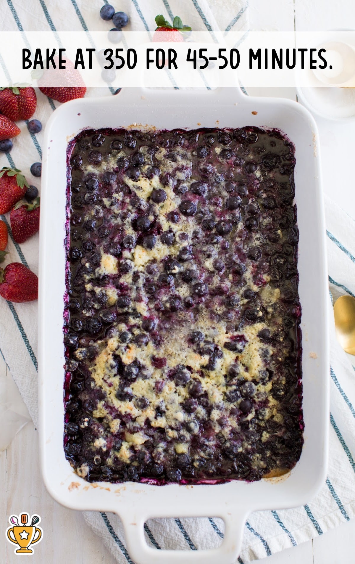 Baked blueberry cobbler in a large white baking dish on a striped towel with strawberries around. Text overlay says "Bake at 350 for 45-50 minutes."