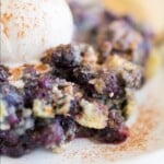 Close-up of a serving of blueberry cobbler with golden crust and juicy blueberries, topped with a scoop of vanilla ice cream.