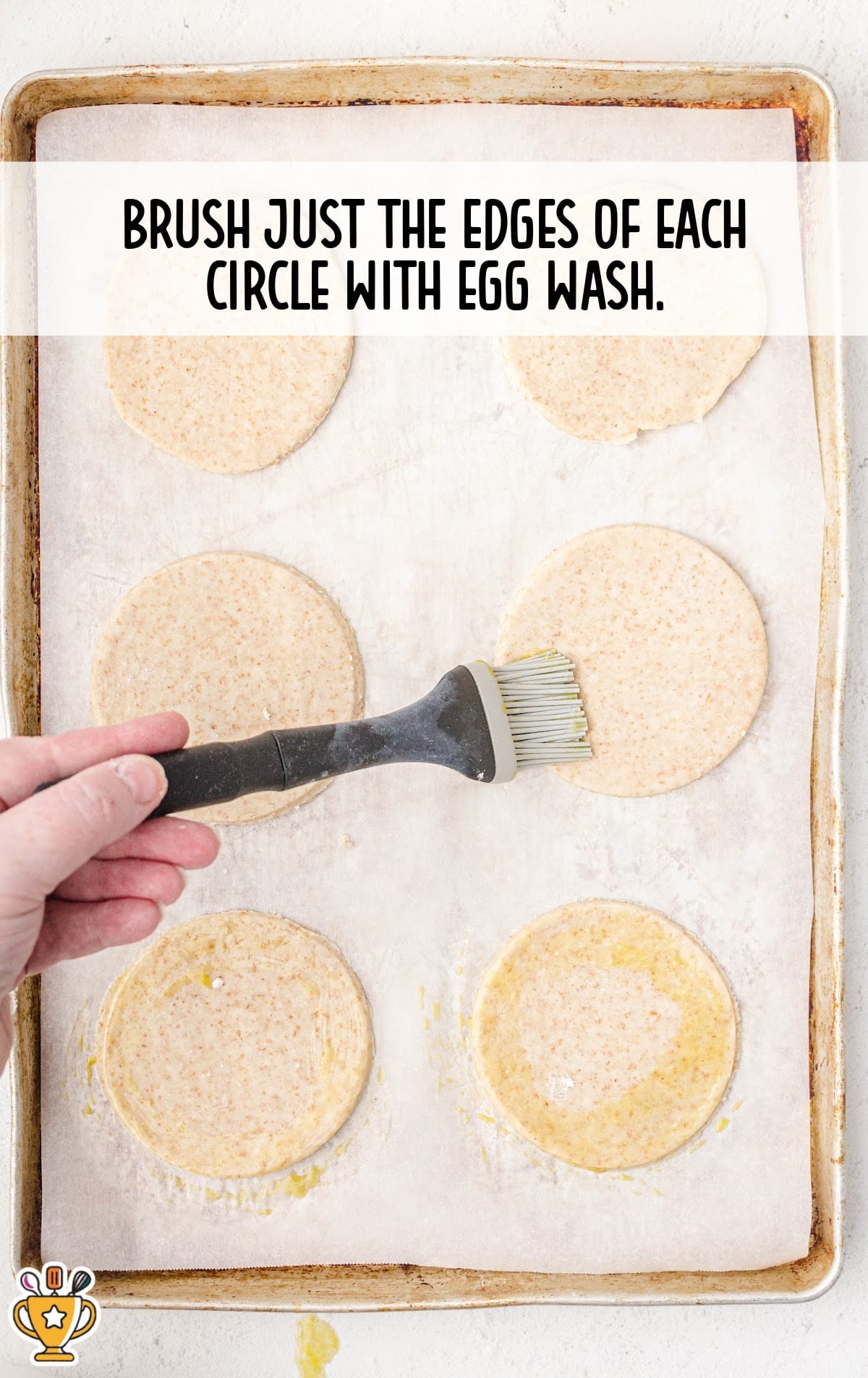 Hand brushing egg wash onto the edges of round pie dough cutouts on a parchment-lined baking sheet.