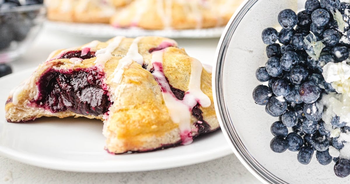 Close-up of a golden-brown blueberry hand pie with white glaze drizzle and a juicy blueberry filling, alongside a bowl of fresh blueberries mixed with sugar and lemon zest