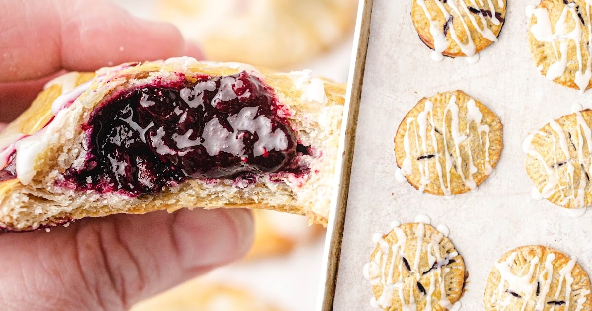 A hand holding a blueberry hand pie with a vibrant jam-like filling, paired with a baking tray full of baked, round hand pies drizzled with icing.