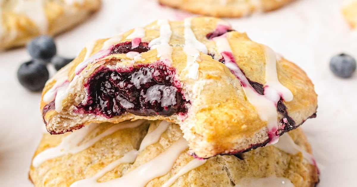 Stacked blueberry hand pies with flaky, golden crusts and sweet blueberry filling, topped with white icing and surrounded by fresh blueberries.