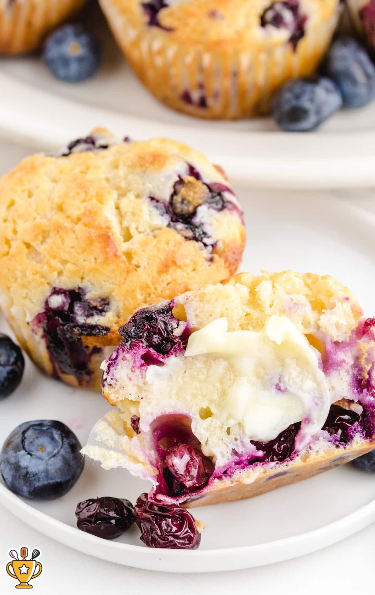 Butter-melted blueberry muffin cut in half, surrounded by whole muffins and fresh blueberries on a white plate.