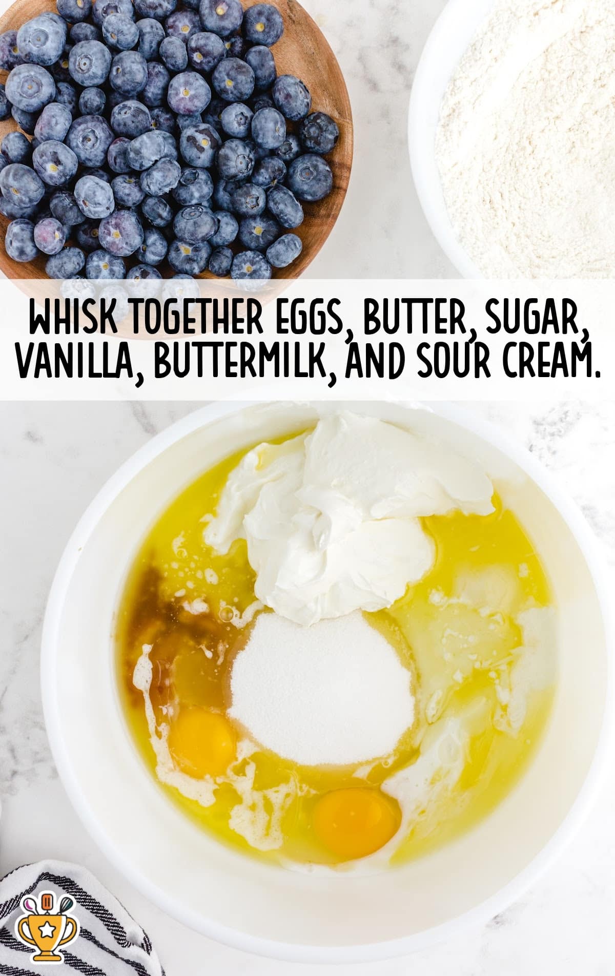 Bowl of eggs, sour cream, sugar, butter, and vanilla ready to be whisked together, next to flour and fresh blueberries.