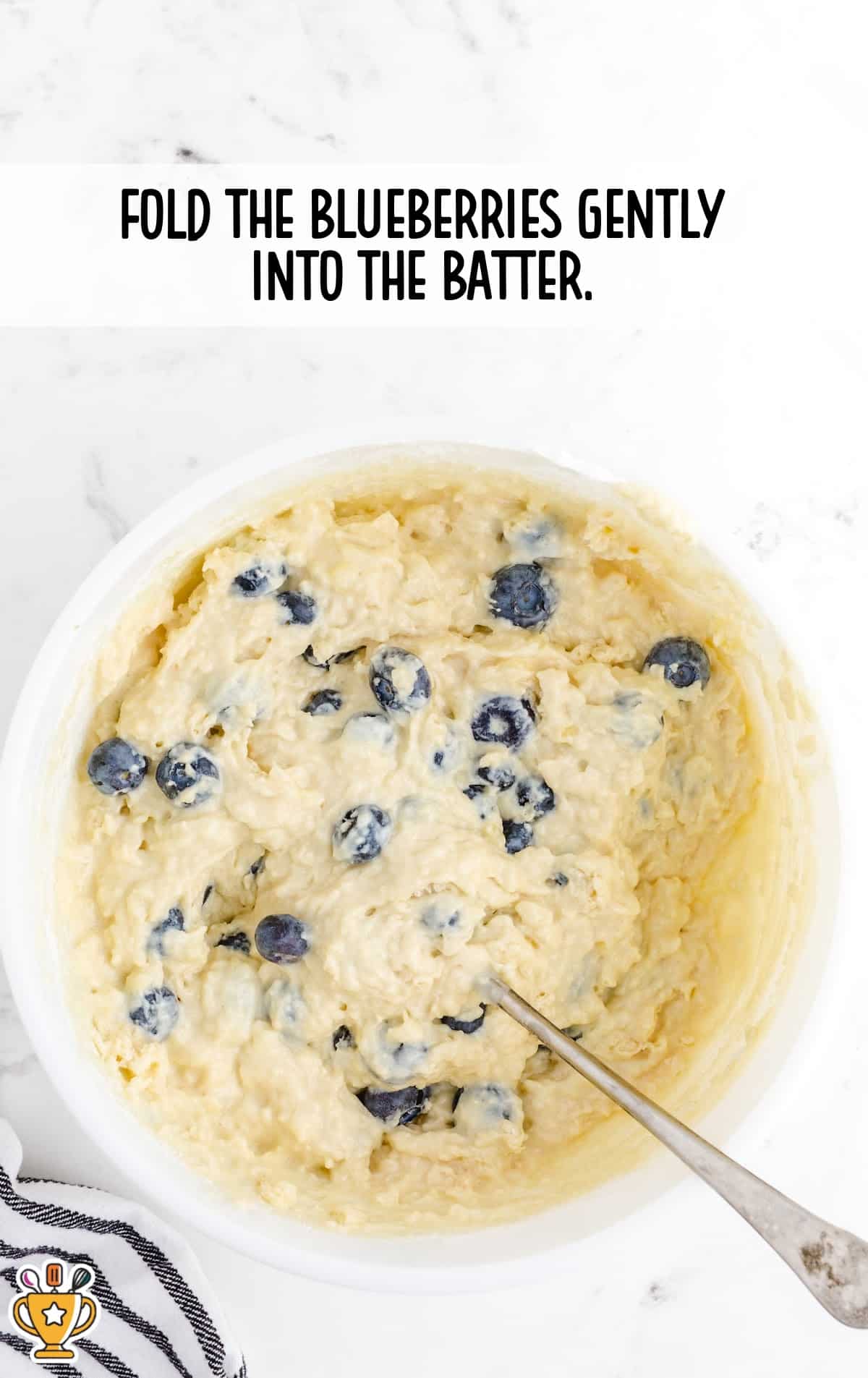 Fluffy batter filled with plump blueberries being stirred gently in a white mixing bowl.