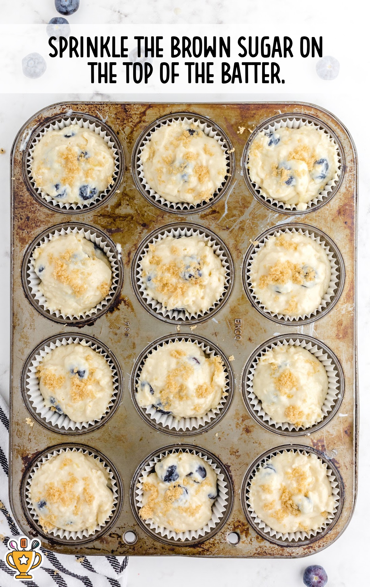 Muffin tin filled with blueberry batter topped with a sprinkle of light brown sugar before baking.