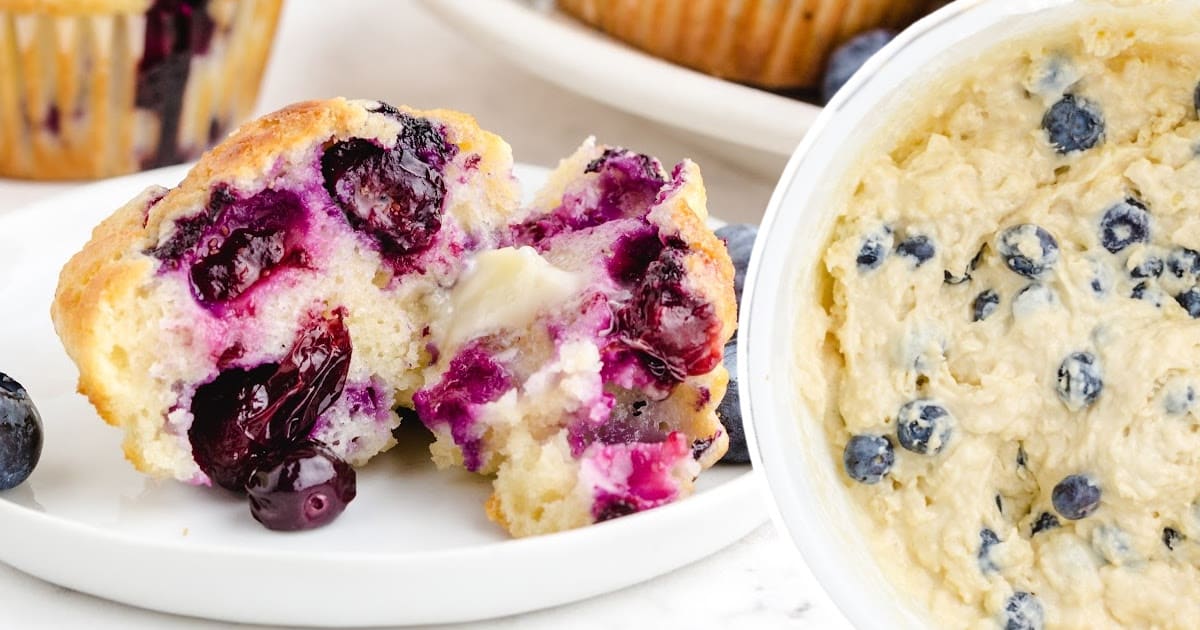 Blueberry muffin split in half showing its gooey center, placed next to a bowl of thick blueberry batter.