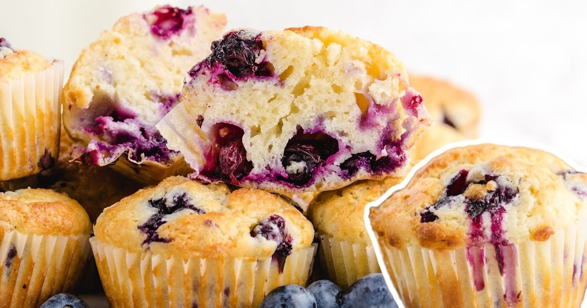 Pile of blueberry muffins with one halved muffin on top showcasing juicy blueberries and fluffy texture.