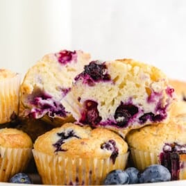 Blueberry muffins stacked on a plate, one muffin halved with juicy berries inside and surrounded by whole blueberries.