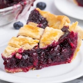 Slice of homemade blueberry pie on a white plate with fresh blueberries scattered around, showcasing the juicy berry filling.