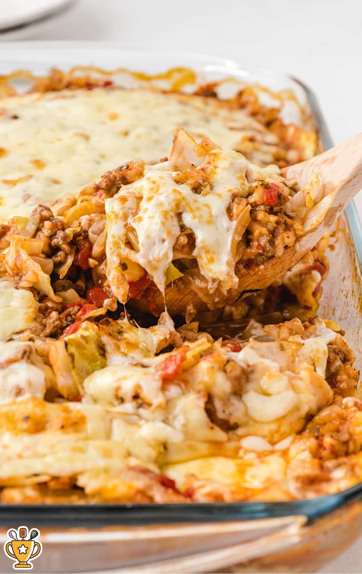Glass baking dish filled with melted cheesy cabbage roll casserole being served with a wooden spoon.