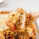 Wooden spoon lifting a cheesy serving of cabbage roll casserole with ground beef, rice, tomatoes, and cabbage from a baking dish.