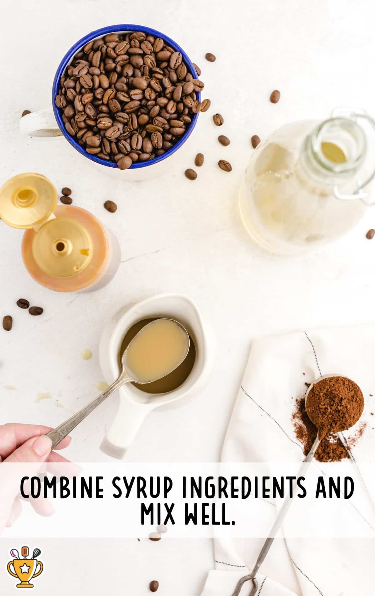 Hand holding a spoon above a small pitcher of caramel mixture, surrounded by coffee syrup ingredients on a white countertop.