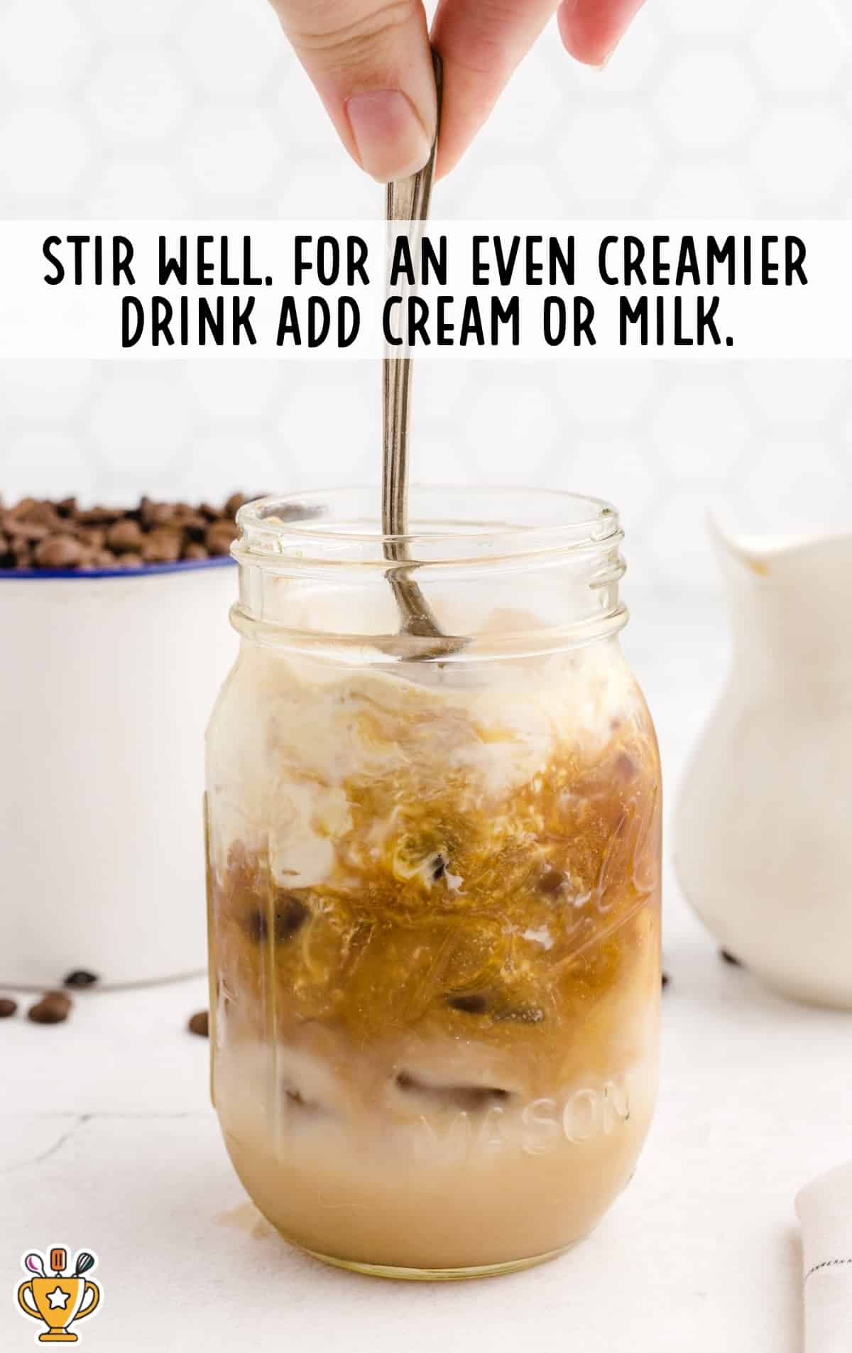 Hand stirring a glass of iced caramel macchiato with cream swirling through the coffee mixture in a mason jar.
