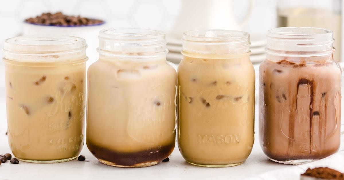Four iced caramel macchiatos lined up in mason jars, each in different tones from light to dark coffee color, on a white surface.