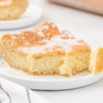 Wooden spatula lifting a cheesy danish slice with melty icing from a glass dish. Casserole shows golden, flaky top.