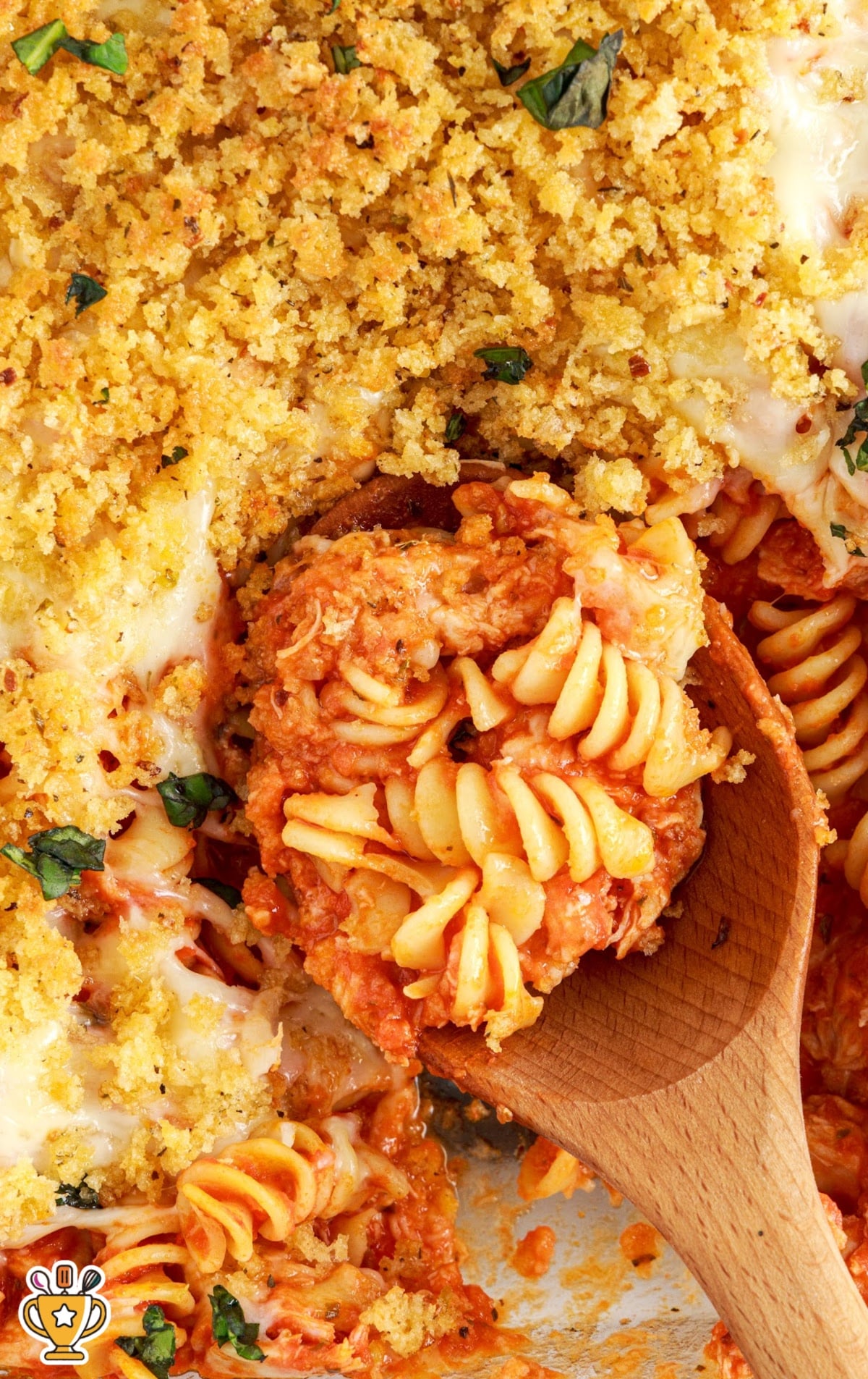 Overhead view of a casserole dish filled with Chicken Parmesan Casserole, a wooden spoon lifting a portion of pasta, chicken, cheese, and breadcrumbs from the dish.