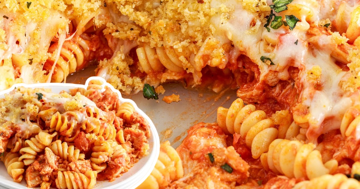Partial casserole dish with a large scoop of Chicken Parmesan Casserole removed, exposing gooey cheese, marinara-soaked pasta, and toasted breadcrumb topping with chopped basil.