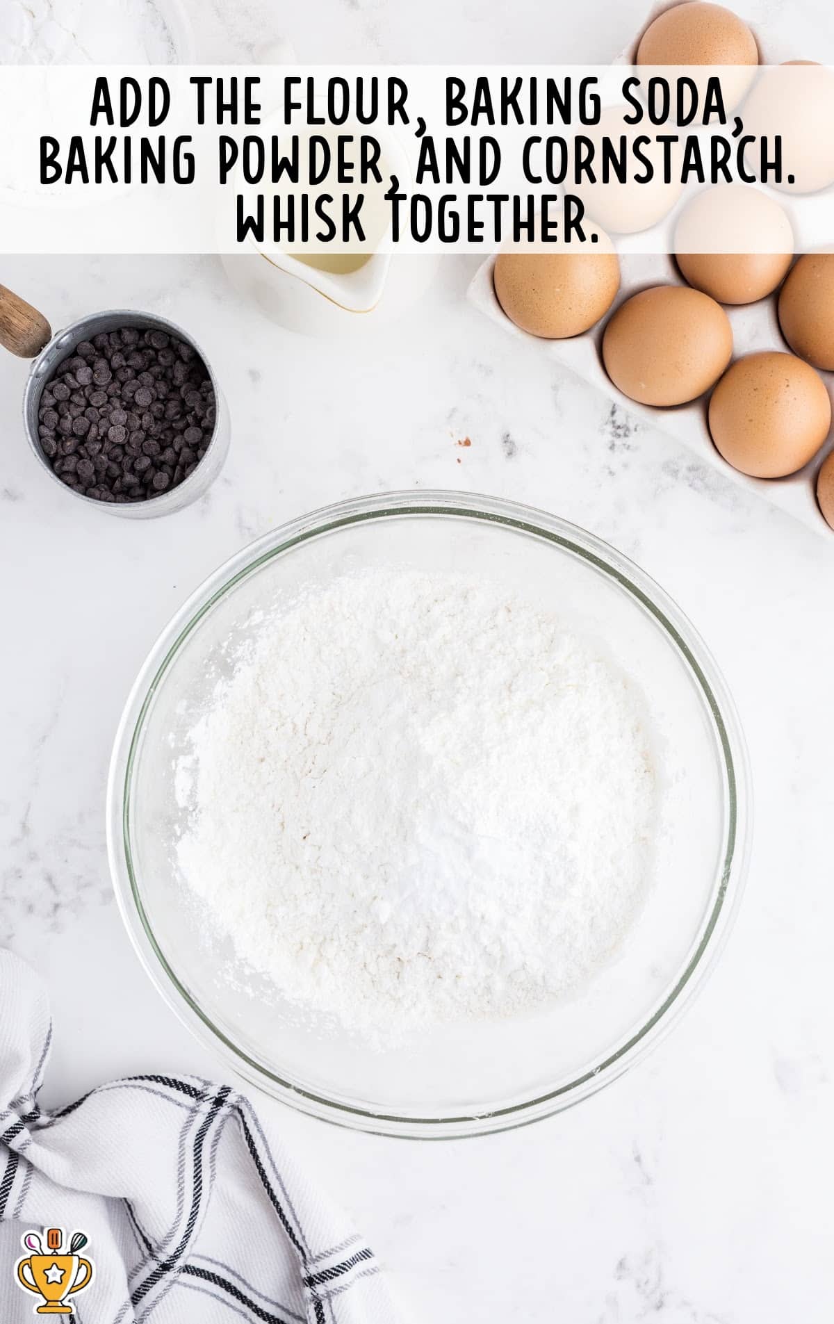 Glass bowl filled with flour, baking powder, baking soda, and cornstarch next to eggs and chocolate chips on marble counter.
