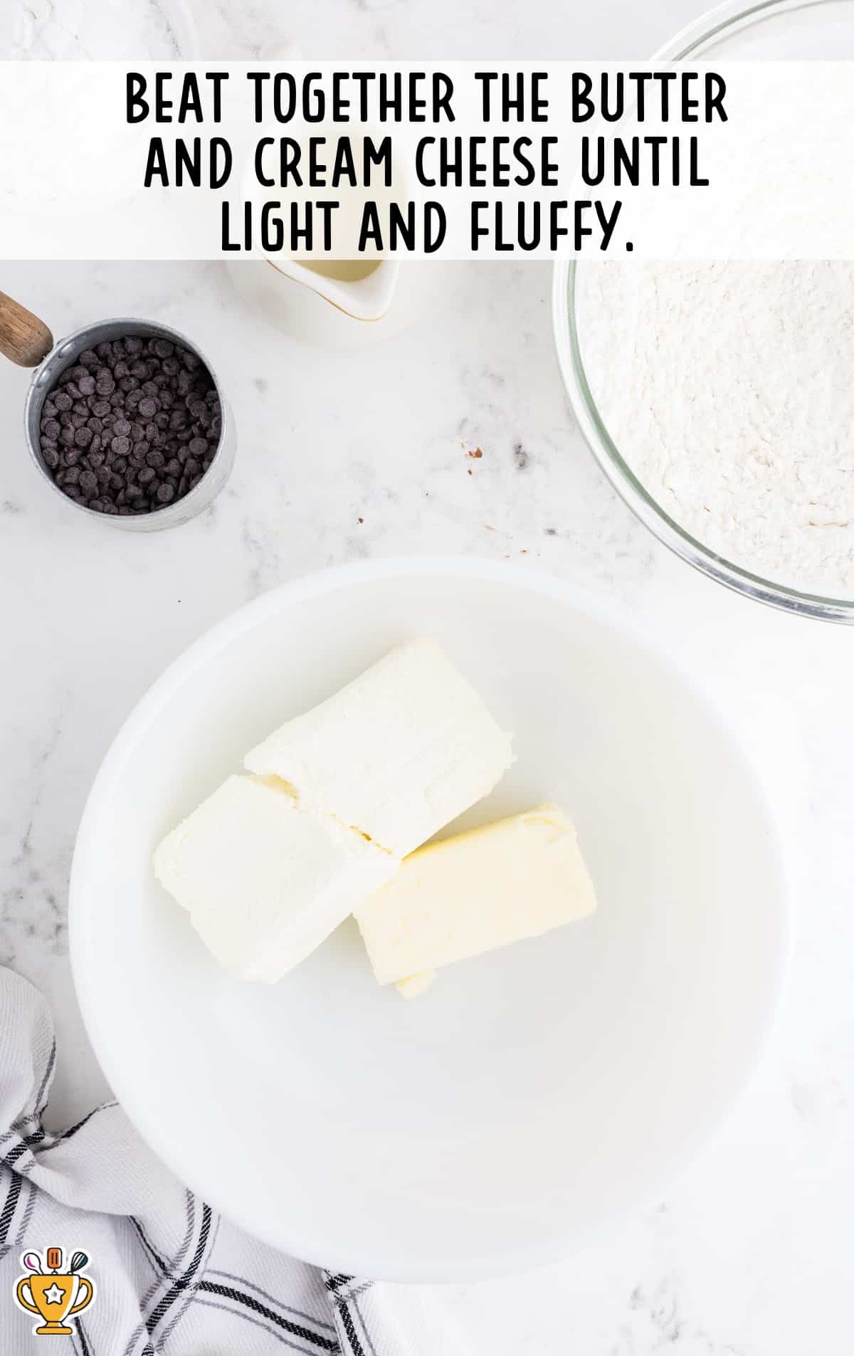 Butter and cream cheese placed in a white mixing bowl on a marble countertop with baking ingredients around.