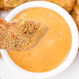Hand holding a crispy chicken tender dipped in creamy copycat Chick-fil-A sauce with more tenders and sauce bowl in the background.