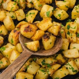 Cast iron skillet filled with golden fried potatoes, being scooped with a wooden spoon and garnished with fresh parsley.