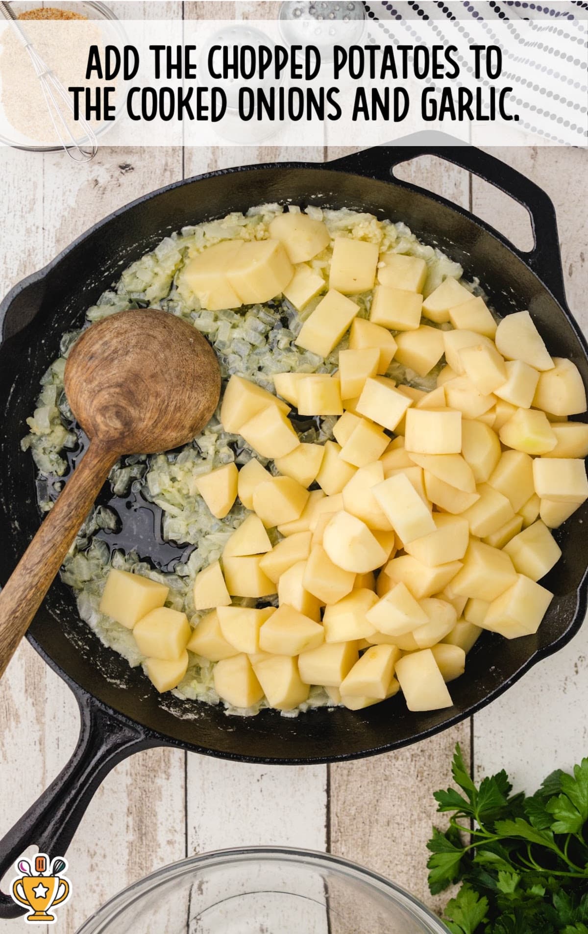 Chopped potatoes added to a skillet with saut&eacute;ed onions and garlic, being stirred with a wooden spoon over a white rustic surface.