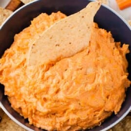Bowl of creamy buffalo chicken dip surrounded by tortilla chips and sliced vegetables on a wooden tray.