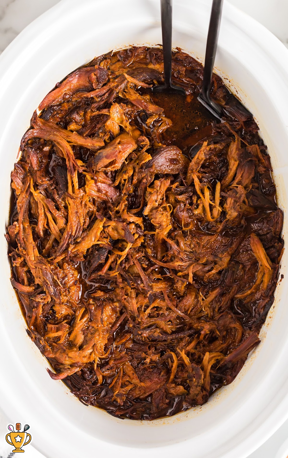 Close-up of shredded Dr. Pepper pulled pork in a white crockpot with two serving forks nestled in the meat.
