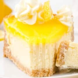 A creamy slice of lemon cheesecake with lemon curd topping, whipped cream swirls, and a graham cracker crust on a white plate.