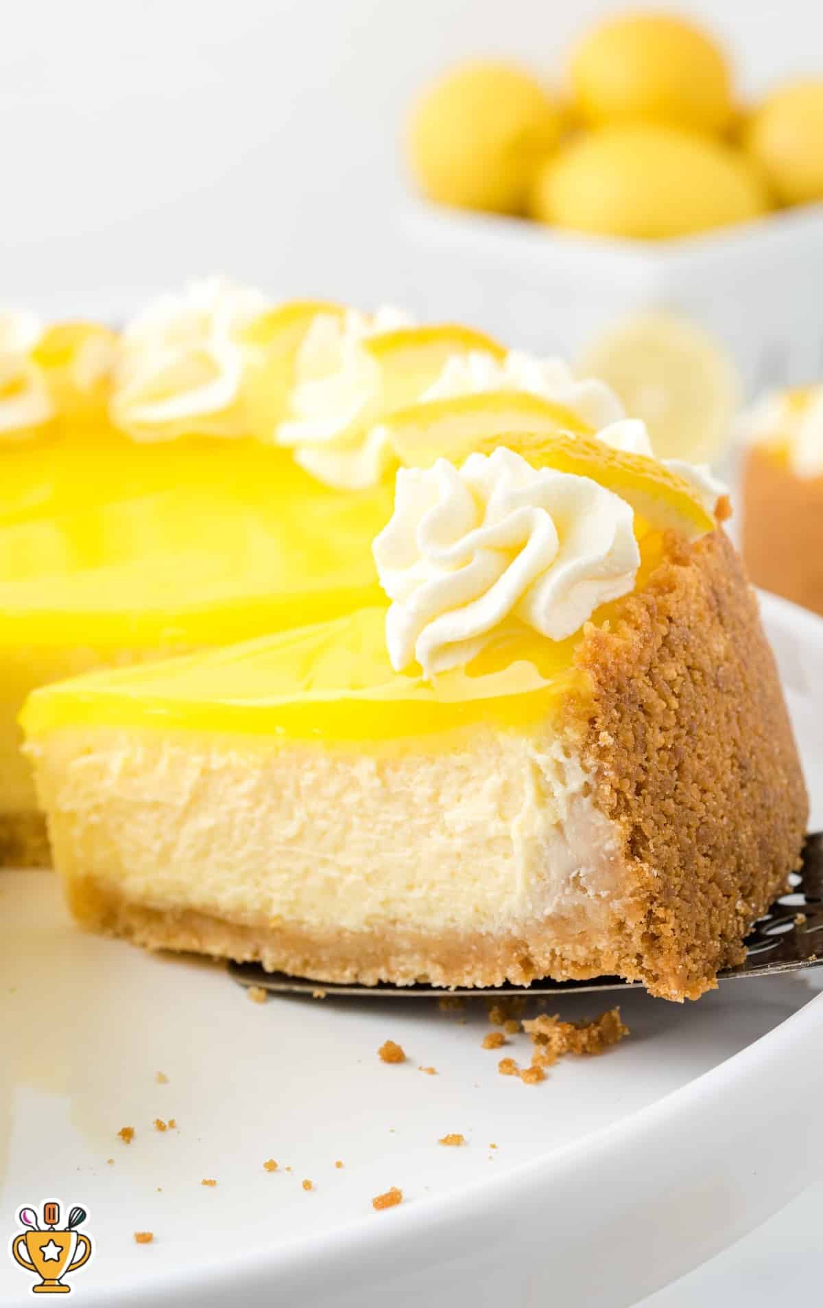 A close-up of a slice of lemon cheesecake with whipped cream swirls and lemon wedge garnish, highlighting the smooth filling and thick crust.