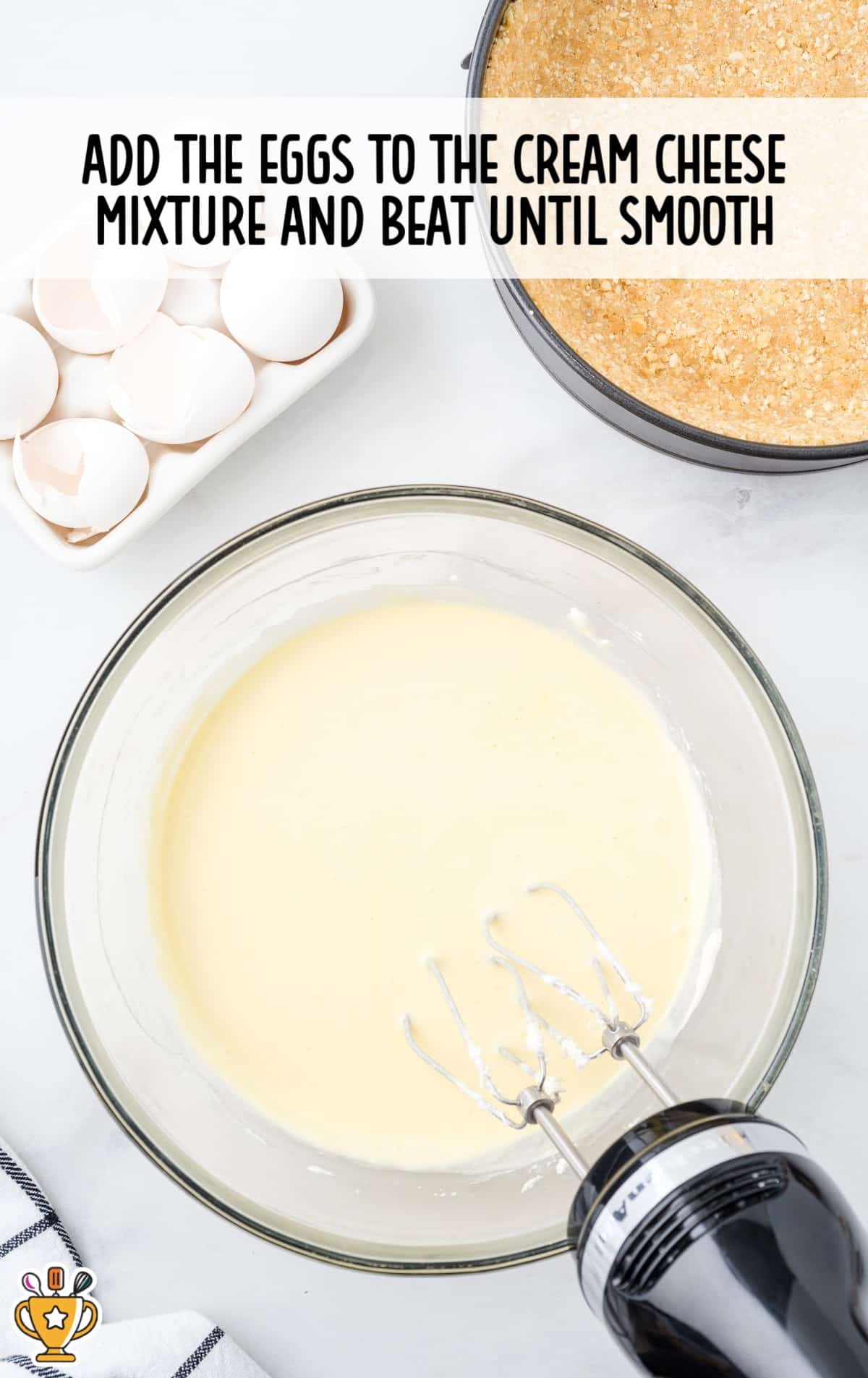 Cream cheese mixture being blended with eggs in a mixing bowl using a hand mixer, with egg shells and crust nearby.
