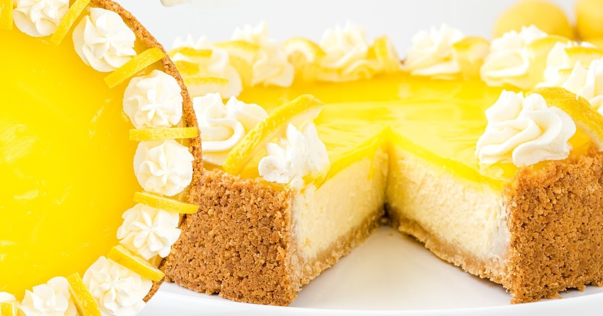 Lemon cheesecake with a slice cut out, showing a creamy interior, piped whipped cream, and fresh lemon slices on top of a golden crust.