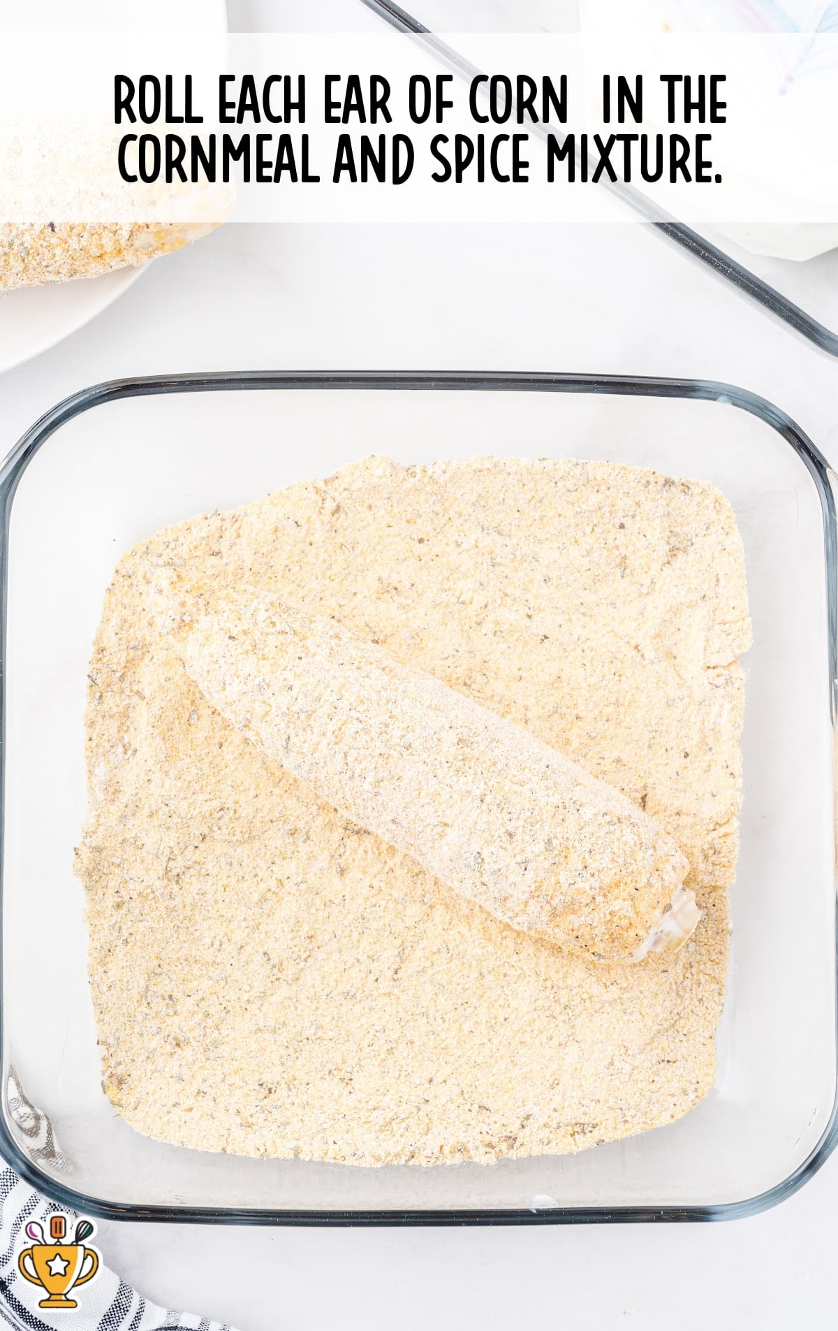 An ear of corn being coated in a cornmeal and spice mixture in a glass dish.