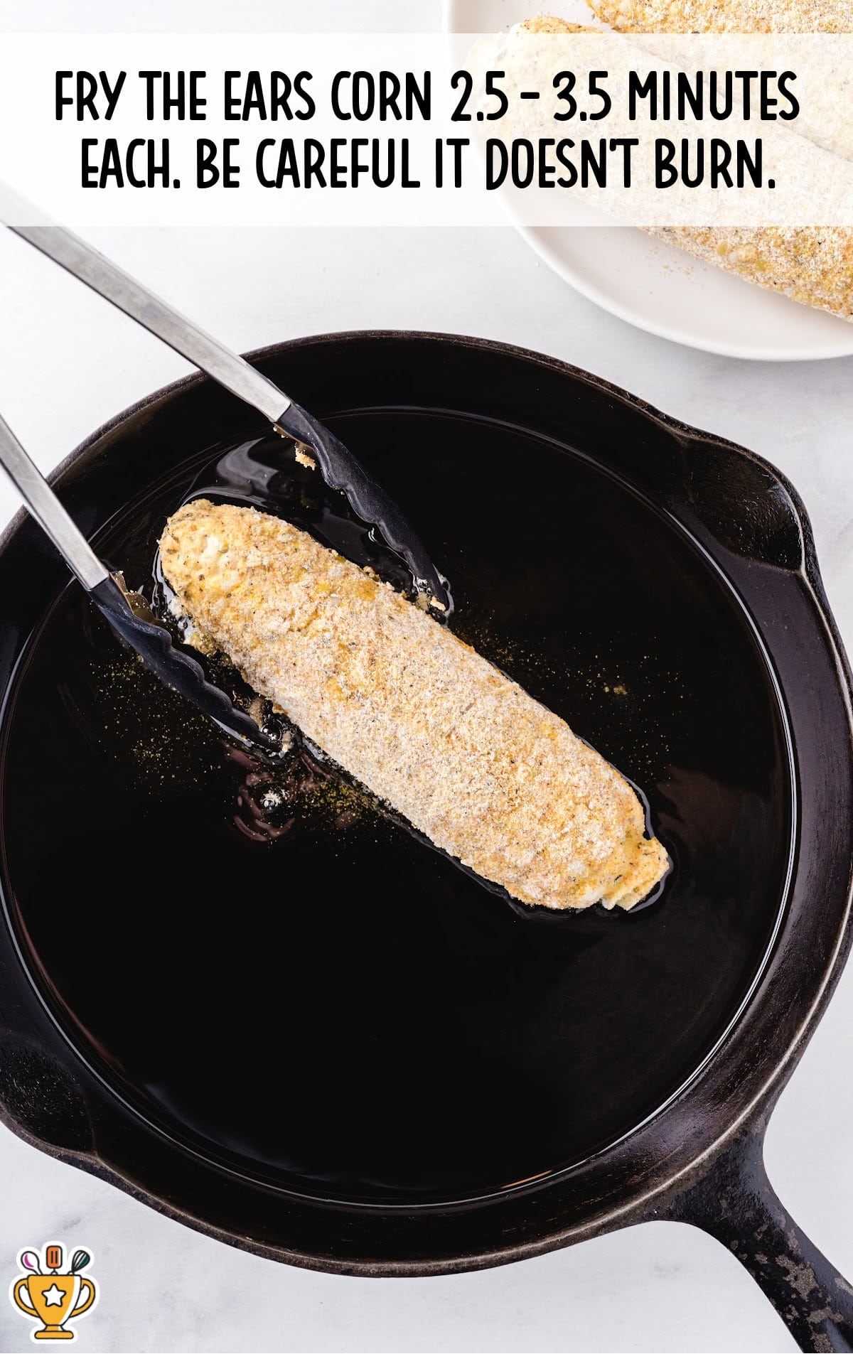 An ear of corn being lowered into hot oil in a skillet with tongs, ready to fry.
