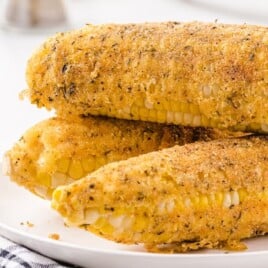 Three pieces of fried corn on the cob stacked on a white plate with a black and white towel nearby.