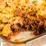Visual recipe card showing labeled ingredients for Frito pie including ground beef, beans, cheeses, salsa, seasonings, and a bag of corn chips, all arranged on a marble background.
