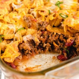 Visual recipe card showing labeled ingredients for Frito pie including ground beef, beans, cheeses, salsa, seasonings, and a bag of corn chips, all arranged on a marble background.