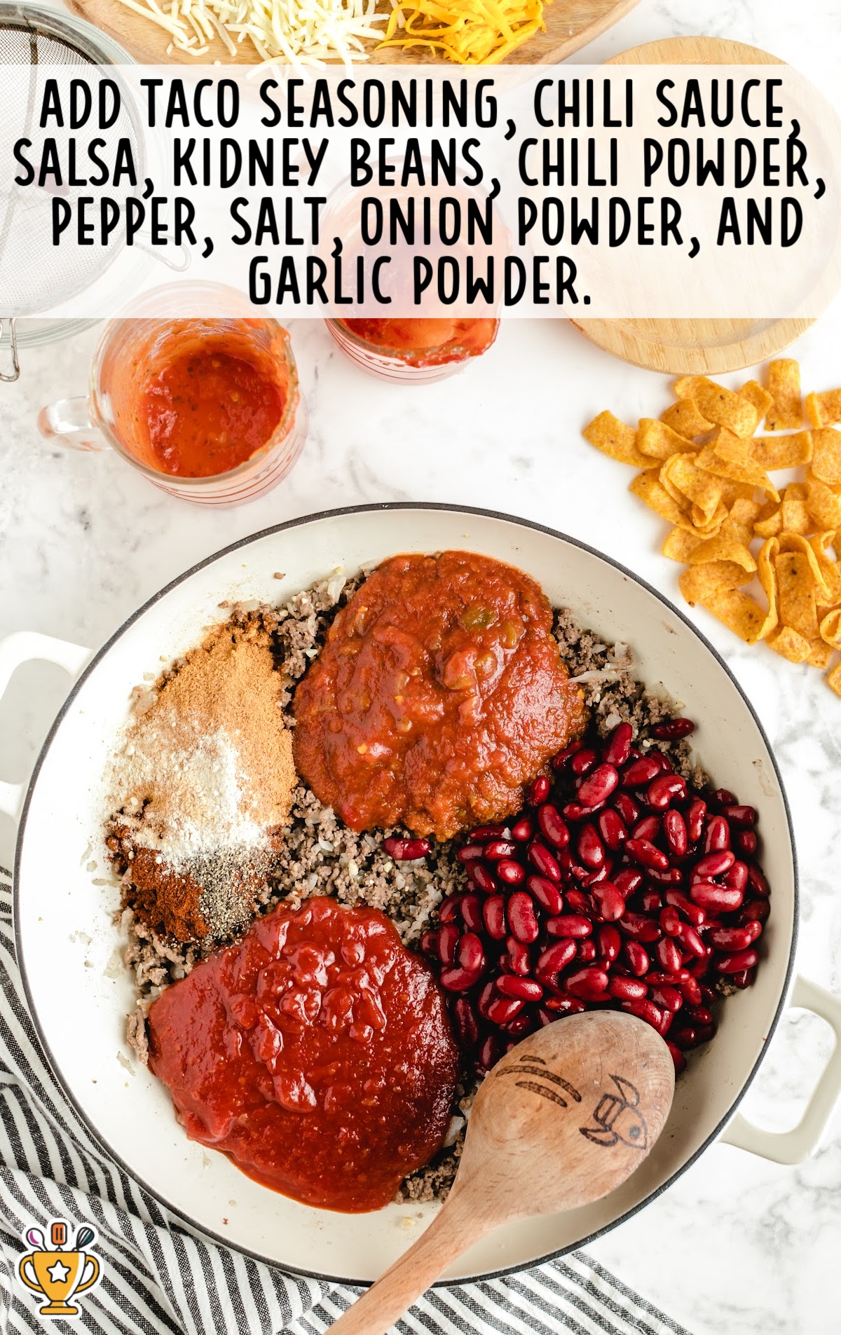 Ground beef mixture in a skillet with kidney beans, salsa, chili sauce, taco seasoning, and various spices spread across the meat, ready to be stirred.