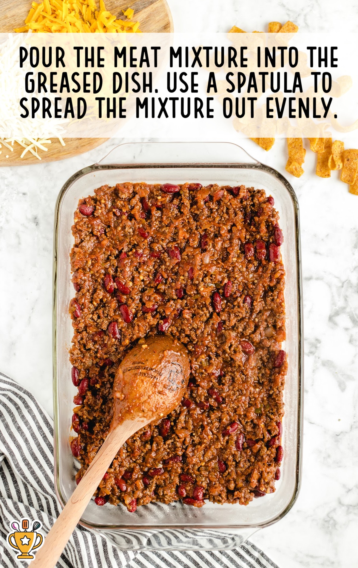 A glass baking dish filled with ground beef and kidney bean mixture spread evenly with a wooden spoon. Cheese and Fritos surround the setup.