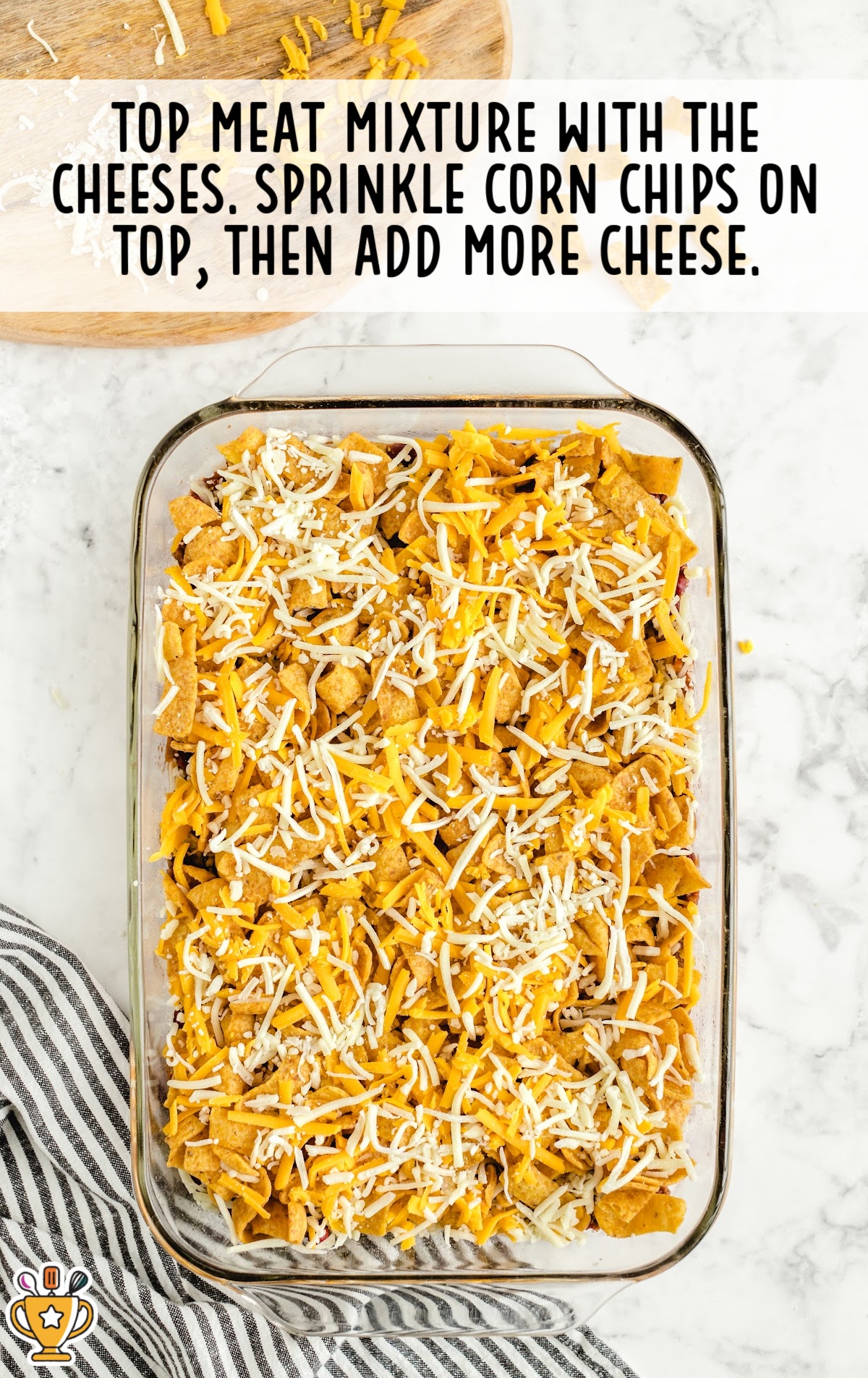 Top-down view of Frito pie being layered&mdash;cheddar and Monterey Jack cheese sprinkled over beef and bean base, followed by corn chips and more cheese.
