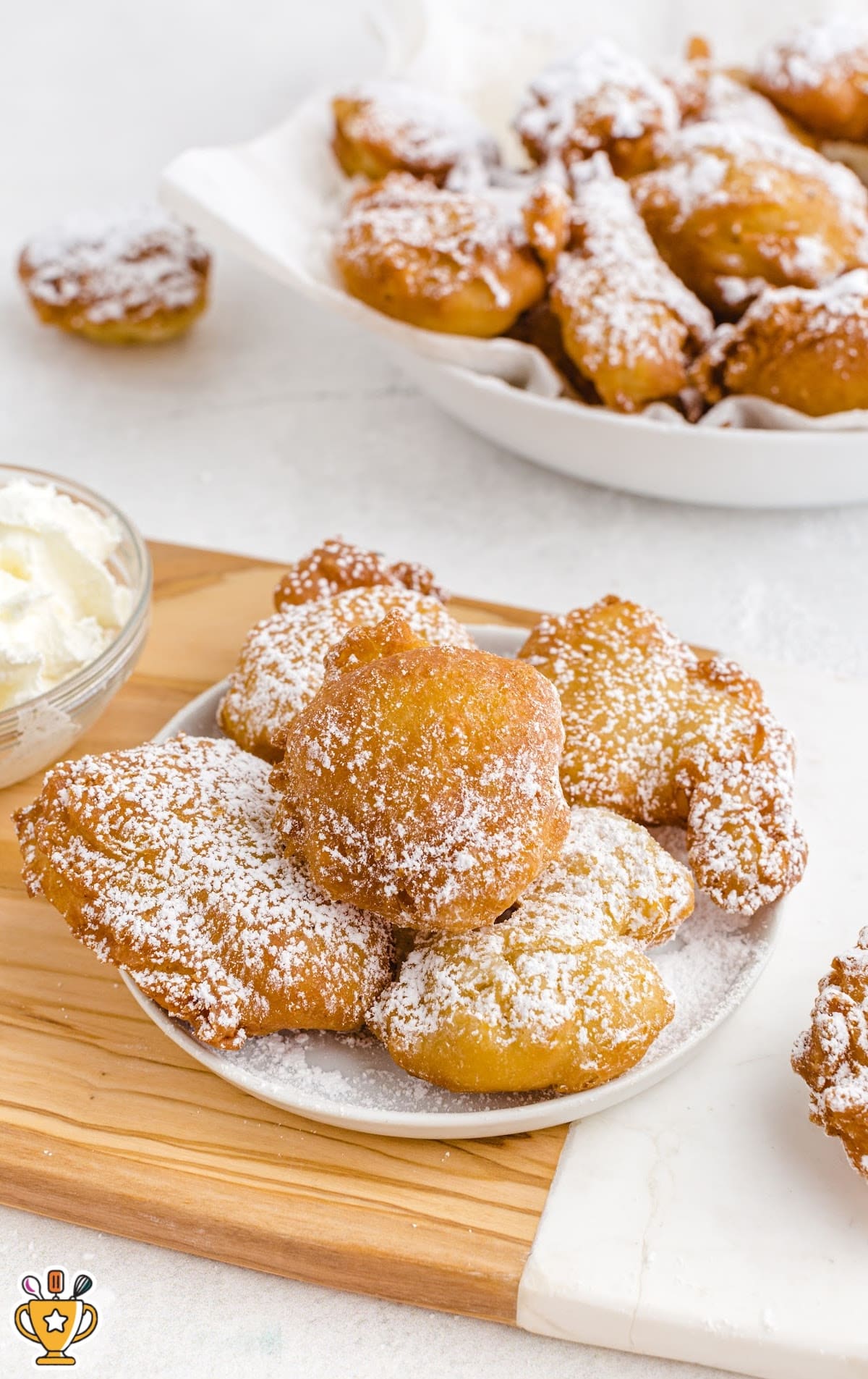Plate of funnel cake bites topped with powdered sugar and served with whipped cream, with more bites in a bowl behind.