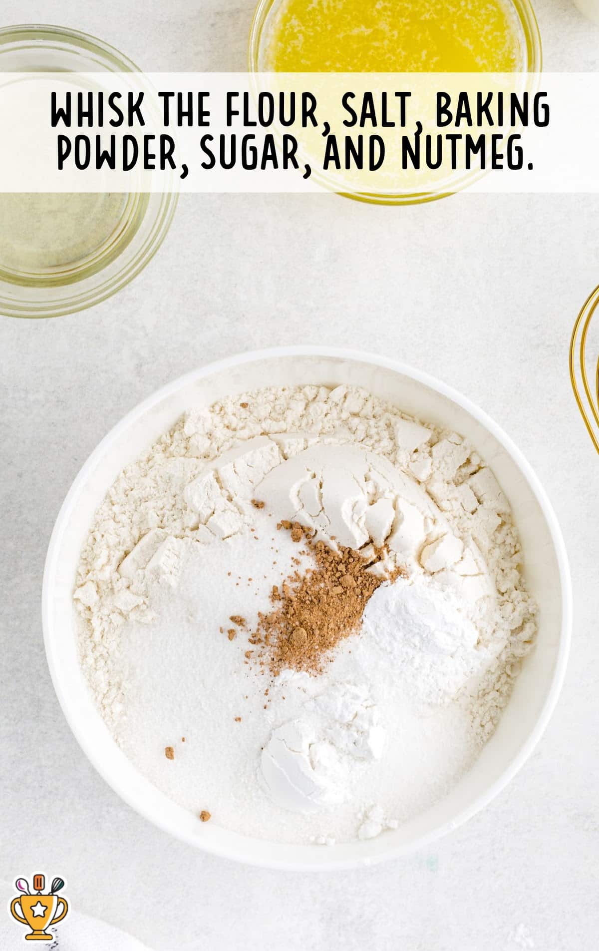 Overhead view of dry ingredients in a white bowl, showing flour, salt, baking powder, sugar, and nutmeg ready to be whisked together.