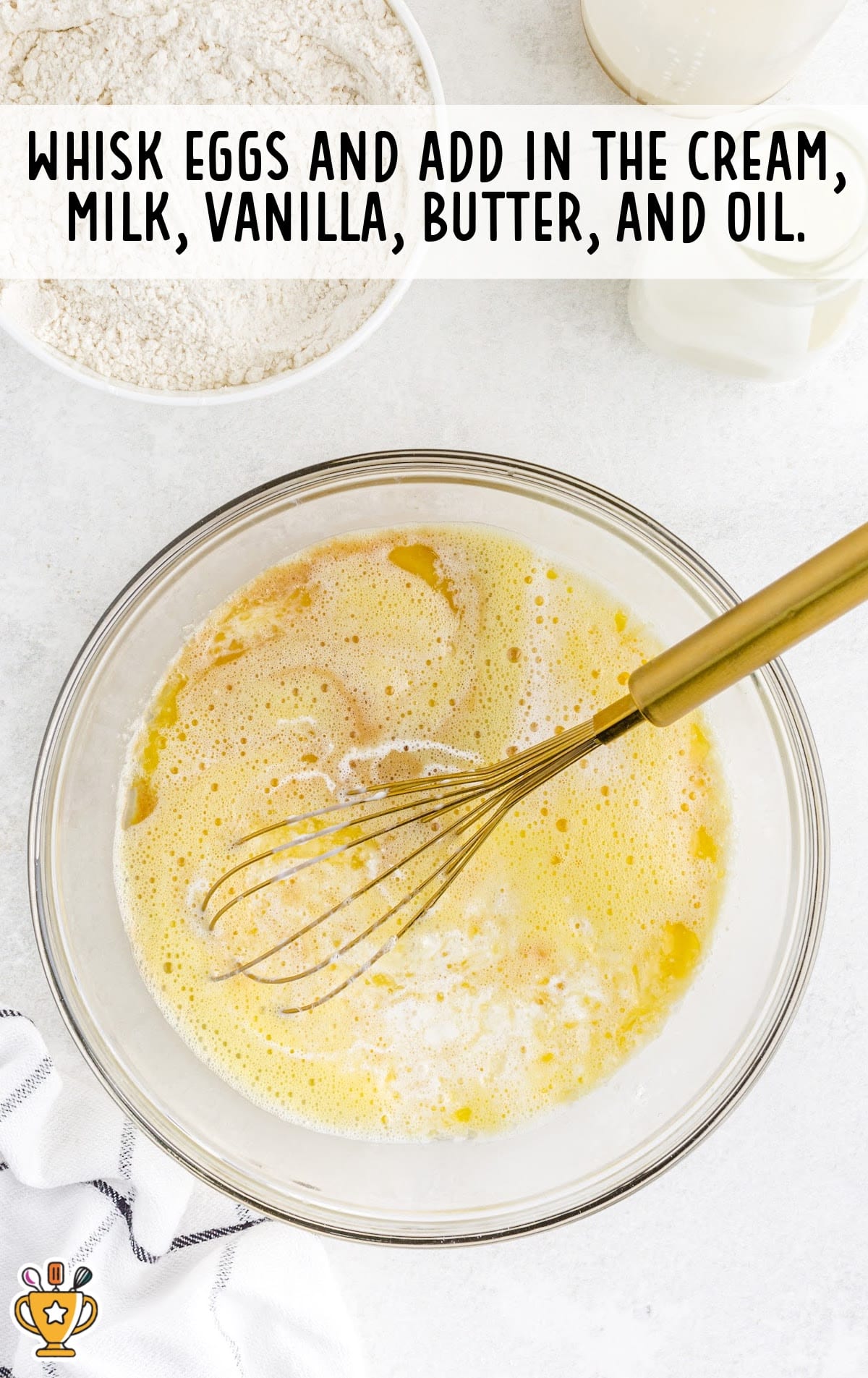 Mixing bowl filled with whisked eggs, cream, milk, butter, vanilla, and oil, with a gold whisk resting inside the wet ingredients.