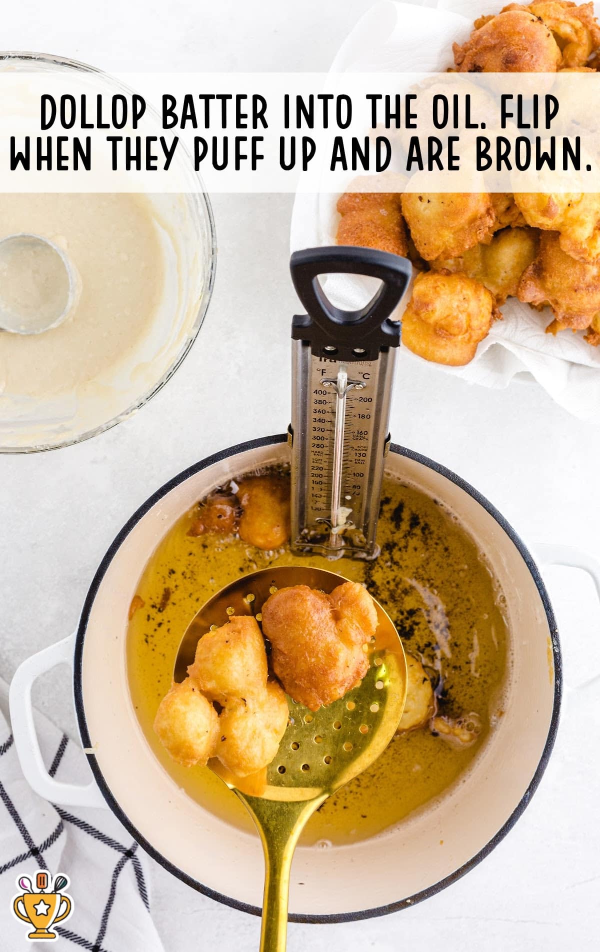 Golden funnel cake bites frying in hot oil, with a thermometer clipped to the pot and a slotted spoon lifting the bites.