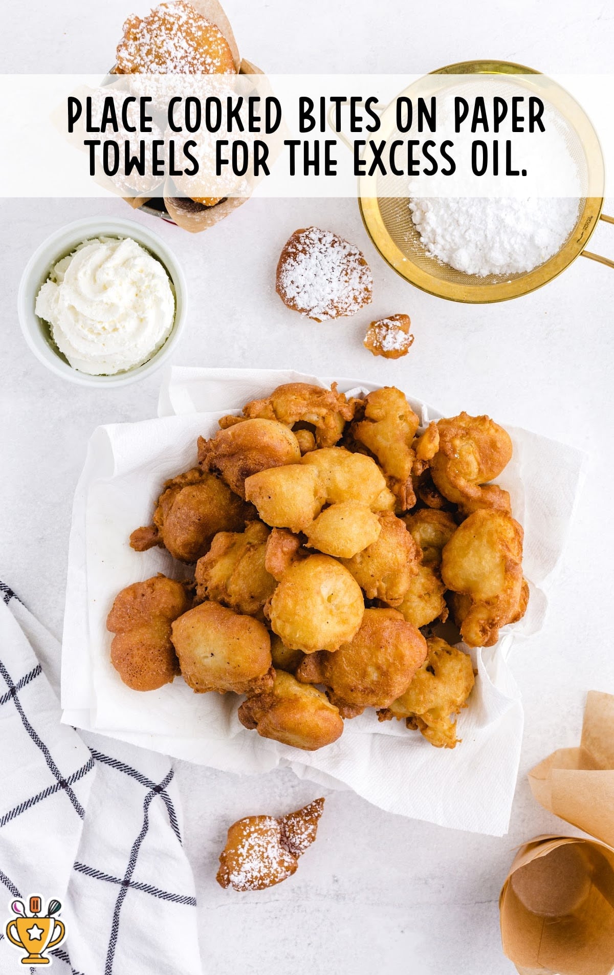 Freshly fried funnel cake bites draining on paper towels with powdered sugar and whipped cream visible in the background.