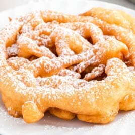 Freshly fried golden brown funnel cake on a white plate, topped with a light sprinkle of powdered sugar.