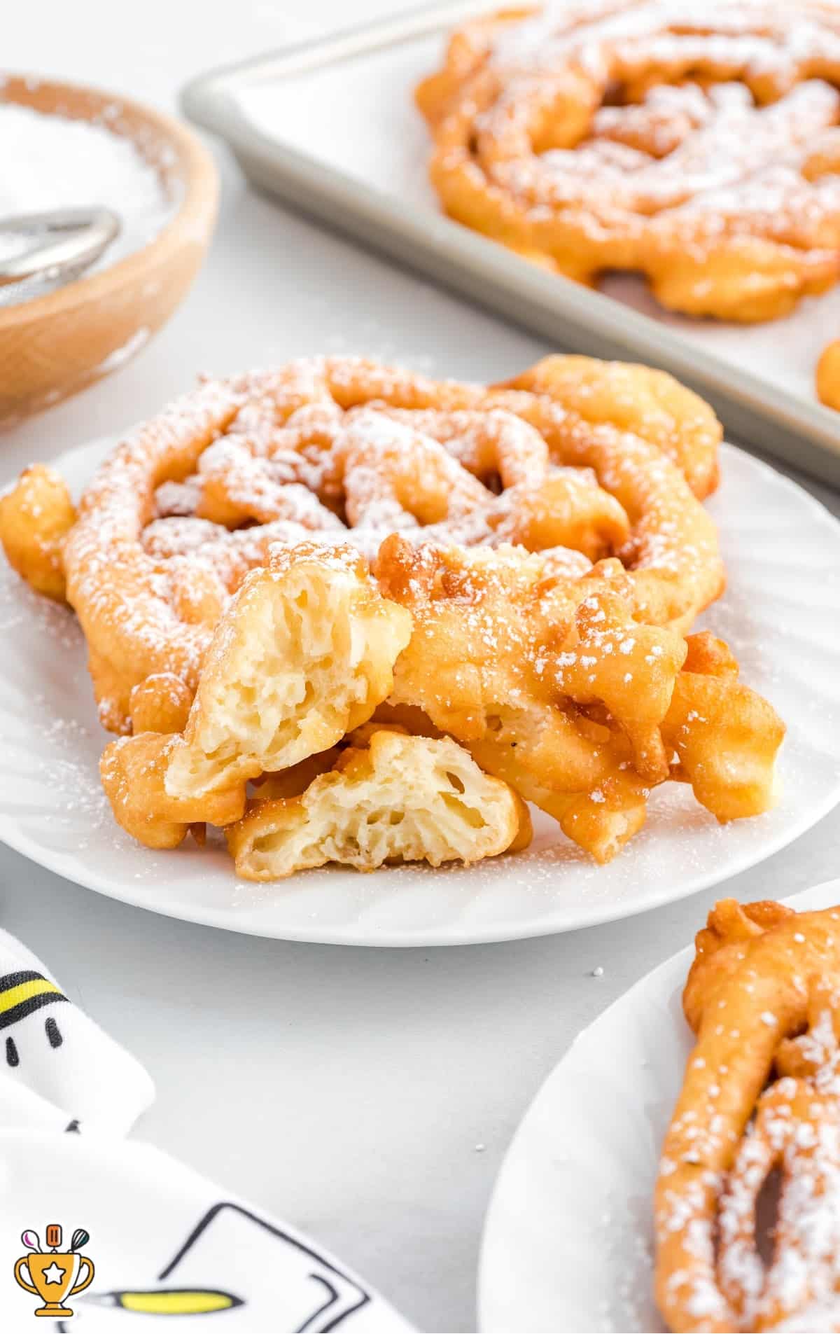 Plate of funnel cake torn in half to show the fluffy, airy texture inside, surrounded by other funnel cakes on the table.